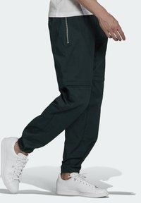 Pantaloni da jogging neri con un design tapered, polsini elasticizzati e un dettaglio della tasca con zip argentata, abbinati a sneaker bianche.
