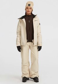 Woman wearing beige winter jacket and pants, brown zip-up underneath, beige beanie, black gloves, and black snow boots, standing facing forward.