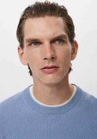 Suéter de canalé azul claro con escote redondeado, superpuesto a una camisa blanca, modelo con cabello corto y ojos azules frente a un fondo liso.