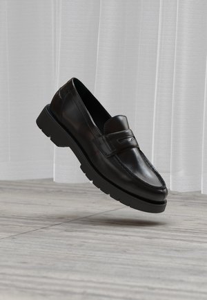 Črni usnjeni loafer z grobo podplatom lebdi nad sivim tlakovcem, v ozadju pa so prosojne bele zavese.