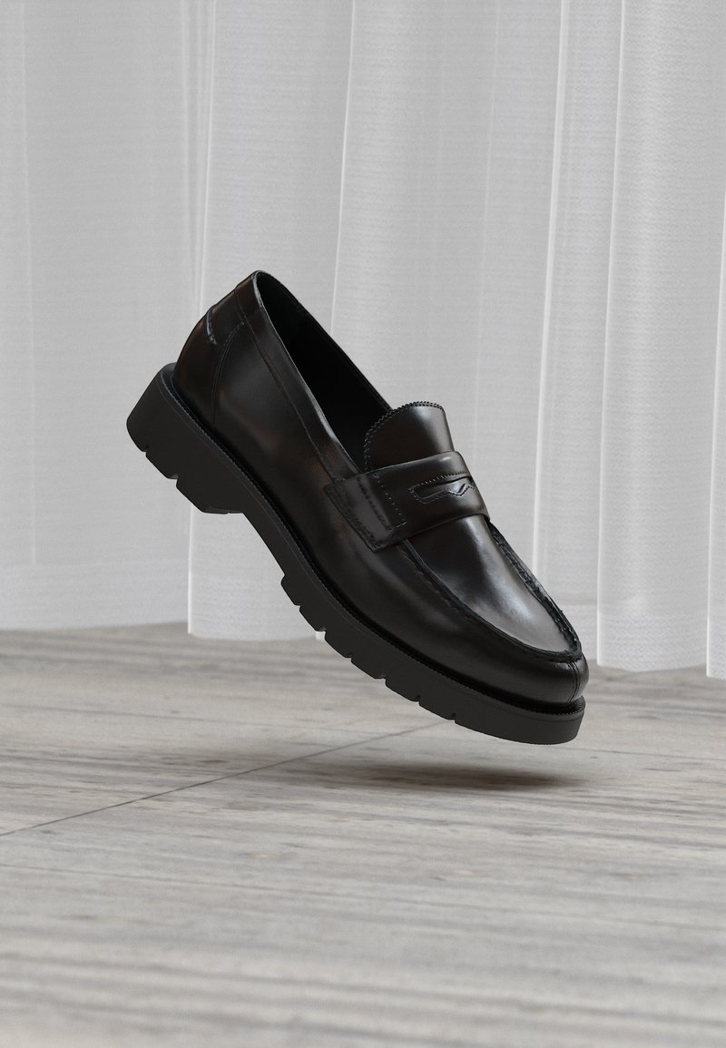 Μαύρο loafer από δέρμα με χοντρή σόλα, που αιωρείται πάνω από γκρι πλακάκια δαπέδου και φόντο από διαφανείς λευκές κουρτίνες.