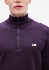 Μωβ πλεκτό πουλόβερ με ψηλό γιακά και μισό φερμουάρ. Διαθέτει το λογότυπο "BOSS" σε λευκό χρώμα στο αριστερό μέρος του στήθους. Απαλή υφή σε όλη την επιφάνεια.