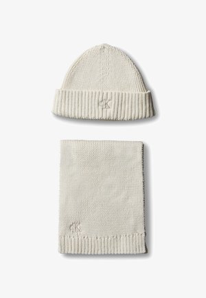 Ensemble bonnet d'hiver en maille crème et écharpe assortie avec bords côtelés et petit logo brodé sur chaque pièce sur fond blanc.