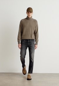 Pulover tricotat de culoare maro cu guler înalt, blugi gri închis, pantofi slip-on bej și o șapcă asortată. Textură netedă și design ajustat pe tot parcursul.