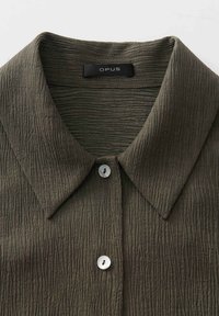 Camicia testurizzata verde oliva con colletto ripiegabile, caratterizzata da una fila di bottoni bianchi e un'etichetta che legge "OPUS."