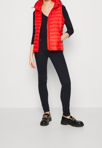 Červená polstrovaná vesta se zapínáním na zip a kapucí, doplněná černými přiléhavými kalhotami a černými botami s detaily zlatého řetízku.