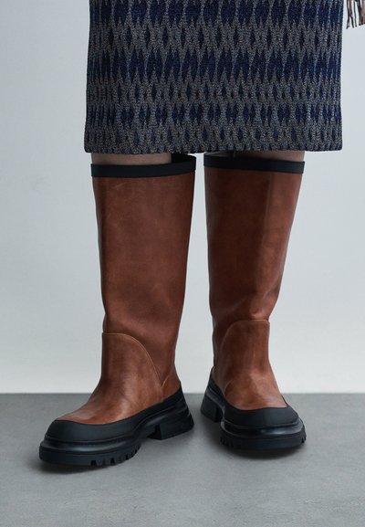 Καφέ δερμάτινες μπότες μέχρι το γόνατο με μαύρες λαστιχένιες σόλες και γείσο. Στρογγυλεμένη μύτη και υφασμένο επάνω μέρος. Συνοδεύονται από φούστα με σχέδιο.