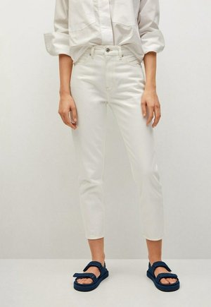 Osoba ubrana w białe krótko obcięte dżinsy, białą koszulę na guziki z podwiniętymi rękawami oraz granatowe sandały z paskami, stojąca na jednolitym tle.