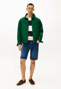 Giacca leggera verde, maglietta a righe blu e bianche, pantaloni corti in denim e scarpe marroni con lacci. Design semplice e casual senza motivi.
