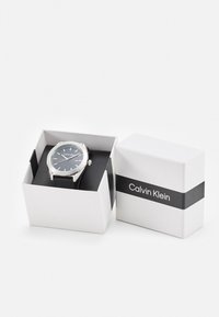 Reloj Calvin Klein con caja de metal plateada, esfera negra y correa de cuero negra, presentado en una caja blanca con una banda negra.