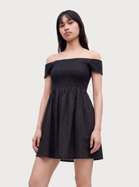Ung kvinde iført en sort off-shoulder kjole med en rynket korsettop og korte ærmer mod en ensfarvet lys baggrund.