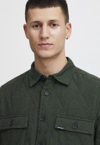 Grønn quiltet jakke med krage, utsmykket med svarte knapper og to brystlommer. Stoffet har en myk tekstur og et subtilt mønster.