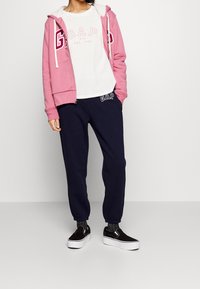 Rosa zip-up hoodie med vit fleecefoder, parat med en vit t-shirt med en rosa logotyp. Marinblå sweatpants med logotypdetalj och svarta slip-on skor.