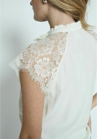 Cremefarvet bluse med blonde detaljer på de korte ærmer, med en flæset halsudskæring og et blødt, let stof.