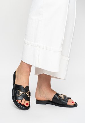 Pieds avec vernis à ongles rouge portant des sandales en cuir noir à boucle dorée, assorties à un pantalon blanc à jambes larges.