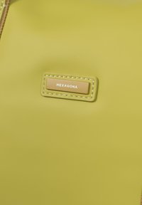 Borsa in materiale verde testurizzato con un'etichetta rettangolare dorata che riporta la scritta "HEXAGONA" e dettagli di cucitura, caratterizzata da un design semplice e moderno.