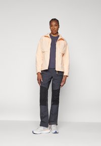 Veste pêche claire avec deux poches avant, t-shirt bleu marine en dessous, pantalon gris foncé avec des sections noires et baskets grises avec des accents orange.