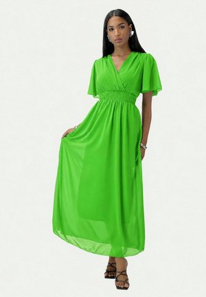 Mujer con el pelo largo y negro que lleva un vestido verde brillante de manga corta hasta los tobillos y sandalias negras de tacón alto con tiras, sujetando el dobladillo del vestido.