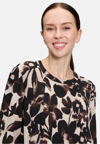 Blusa con stampa floreale in crema, nero e marrone. Presenta delle pieghe al collo e maniche a sbuffo, realizzata in un tessuto leggero e liscio.