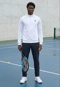 Moški stoji na teniškem igrišču in drži teniško lopar, oblečen je v belo jakno z zadrgo, črne hlače in bele superge.