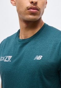 Close-up af en person iført en teal New Balance sportstrøje med "tcs 26" trykt på brystet mod en ensfarvet baggrund.