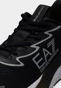 Μαύρο αθλητικό παπούτσι EA7 Emporio Armani με μαύρα κορδόνια, αντανακλαστική ασημένια κυματιστή λεπτομέρεια και λογότυπο στο πλάι και στη γλωττίδα.