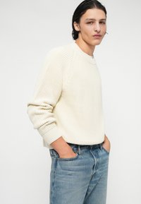 Suéter de punto color crema con textura acanalada y cuello redondo, combinado con jeans de mezclilla azul claro, con las manos metidas en los bolsillos.