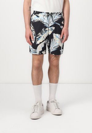 Zwarte zwembroek met een wit en lichtblauw tropisch bladenpatroon, elastische tailleband en zijzakken, gecombineerd met witte sneakers en sokken.