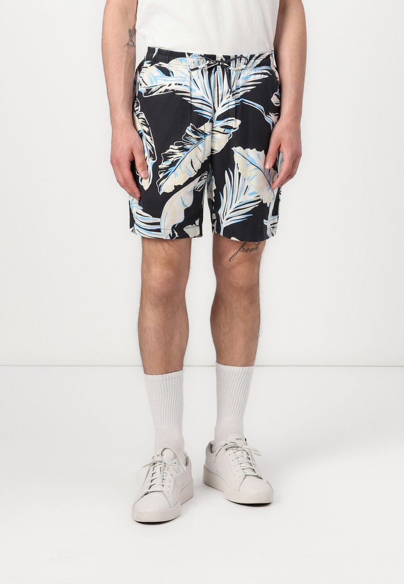 Costumi da bagno neri con motivo tropicale di foglie bianche e azzurro chiaro, vita elastica e tasche laterali, abbinati a sneaker bianche e calze.