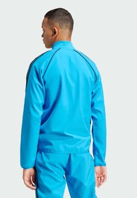 Giacca atletica azzurro chiaro con accenti a strisce nere, tessuto liscio, colletto alto e design aderente. Indossata con pantaloni da tuta abbinati.