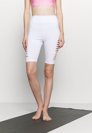 Femme debout pieds nus sur un tapis de yoga noir, portant un short cycliste taille haute blanc avec des découpes latérales et un soutien-gorge de sport rose.