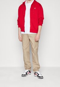 Sudadera roja con cremallera y bolsillo canguro, camiseta blanca, chinos beige y zapatillas negras y rojas con detalles blancos. Atuendo casual.