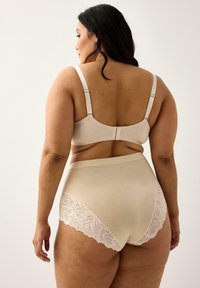 Donna curvy di spalle che indossa un reggiseno beige e mutandine a vita alta con bordo in pizzo su uno sfondo chiaro uniforme.