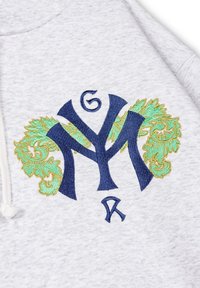 Sudadera gris con un logo bordado en azul oscuro, con intrincados acentos en verde y dorado, sobre un fondo de tela texturizada.