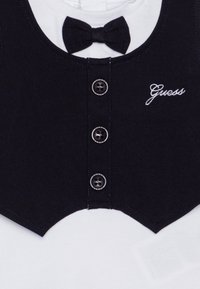 Schwarzes Kragenoberteil mit einem weißen Unterhemd, verfügt über eine Fliege, vier Knöpfe und ein gesticktes "Guess"-Logo in Schreibschrift. Weiche Stofftextur.