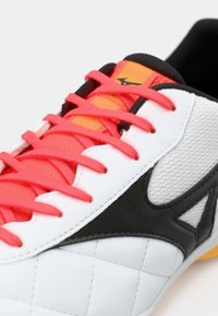 Athletic shoe with white synthetic upper, black accents, and pink laces. Features textured panels and a black padded collar.