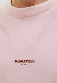 Lyserød bomulds T-shirt med ribbet krave og brun logo tekst "JACK & JONES EST. 1990" foran. Glat tekstur.