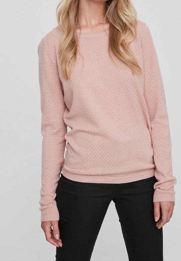 Femme portant un pull texturé rose clair à manches longues et un pantalon noir, debout avec les bras détendus le long du corps.