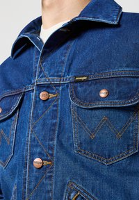 Modrá džínová bunda se klasickým designem, měděnými knoflíky, kontrastním žlutým šitím a dvěma náprsními kapsami s šikmými klopami.