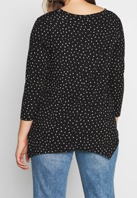 Blusa nera con pois bianchi, maniche a tre quarti, scollatura rotonda e orlo leggermente curvo. Abbinata a jeans blu.
