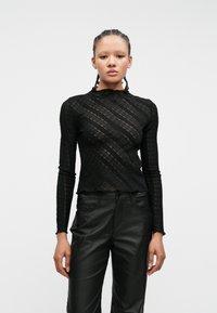 Svart langermet topp med teksturert, mønstrete design og bølget hals, kombinert med svarte skinnbukser.