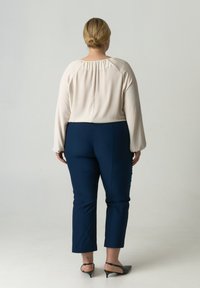 Blouse in lichtroze stof, losse pasvorm met gerimpelde achterkant. Marineblauwe broek, op maat gemaakt, net boven de enkel afgeknipt. Zwarte puntige ballerina's.
