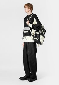 Jersey bicolor negro y crema con detalles plateados, pantalones negros holgados y texturizados, y zapatos negros gruesos. La mochila tiene múltiples bolsillos.