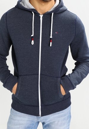 Uomo che indossa una felpa blu navy con zip e cordini bianchi, jeans azzurri, con le mani in tasca, in piedi davanti a uno sfondo semplice.