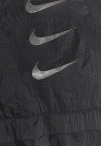 Zwarte stof met een gekreukelde textuur, voorzien van metallic zilveren Nike-logo's die verticaal zijn gerangschikt. Inclusief een naadloze zoom aan de onderkant.