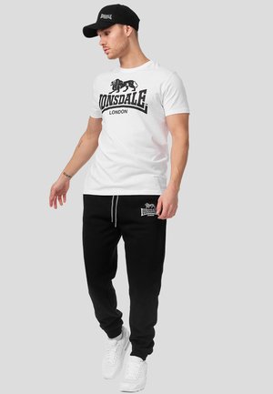 Mand iført hvid Lonsdale London t-shirt, sorte Lonsdale London joggingbukser, hvide sneakers og sort kasket, stående mod en grå baggrund.