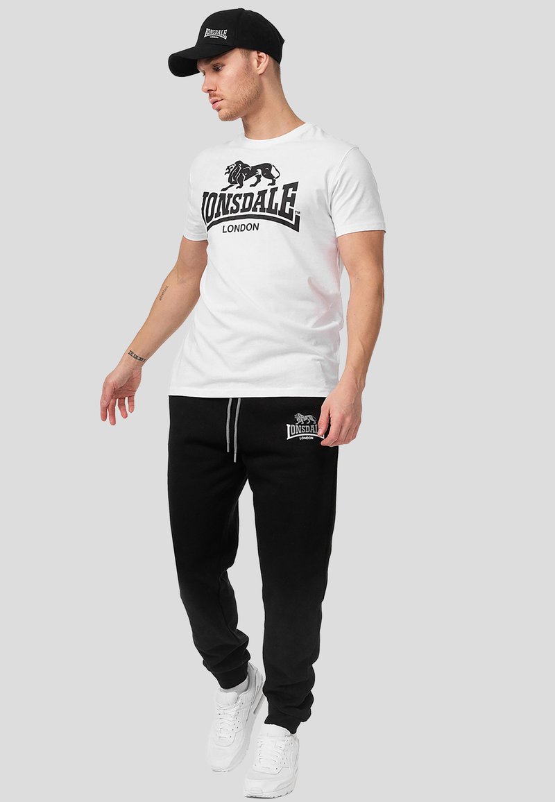 Hombre con camiseta blanca de Lonsdale London, pantalones deportivos negros de Lonsdale London, zapatillas blancas y gorra negra, de pie frente a un fondo gris.