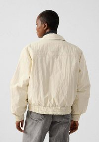 Giacca bomber color crema con tessuto strutturato, vita e polsini elasticizzati, dotata di colletto classico e vestibilità rilassata.