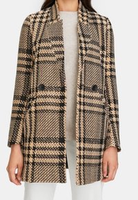 Nainen, jolla on päällään beige ja musta suuri houndstooth-kuvioinen kaksirivinen bleiseri, vaaleanharmaan topin päällä ja valkoiset housut.