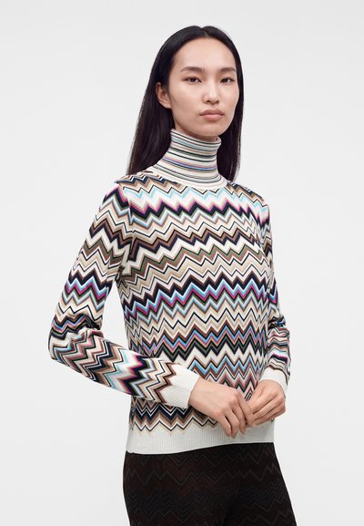 Femme portant un pull à col roulé à manches longues avec un motif zigzag multicolore, debout avec les mains doucement jointes devant elle.